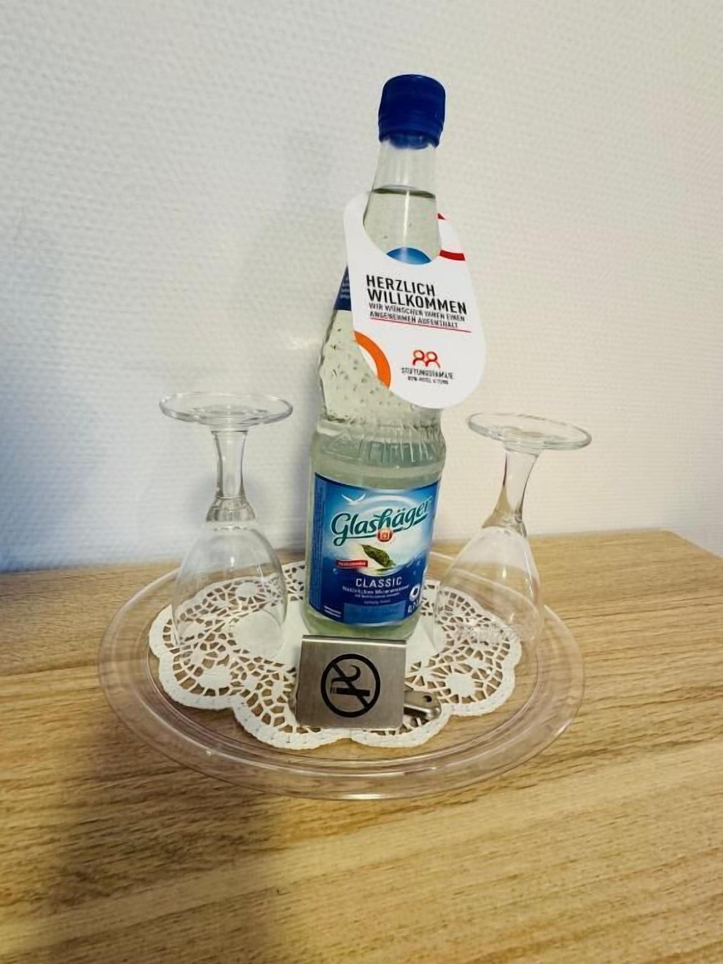 A bottle of Glashäger Classic on a clear glass stand with a doily, surrounded by two empty wine glasses.