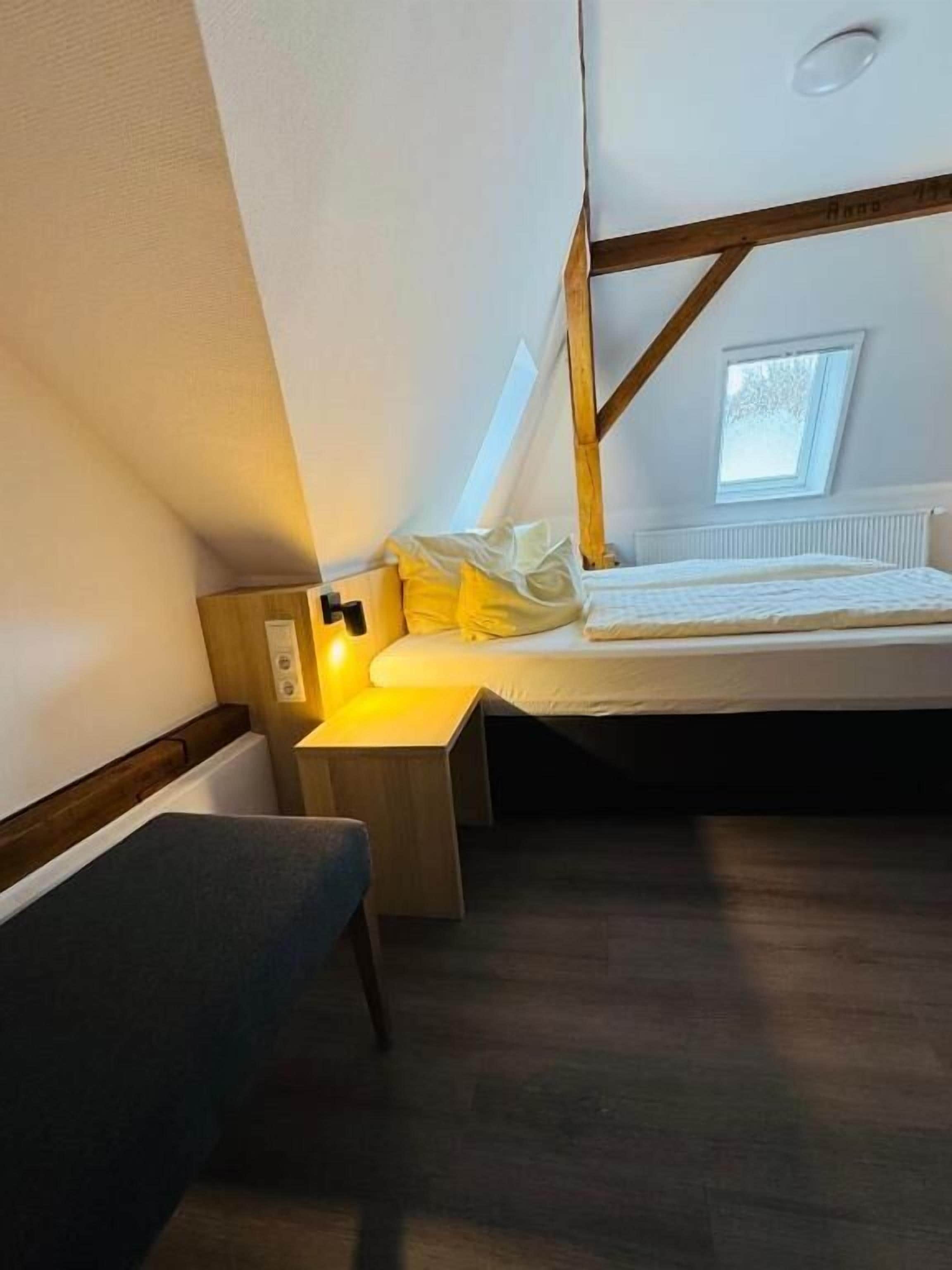 A small, cozy room with a bed, a bench, a nightstand, and a window.