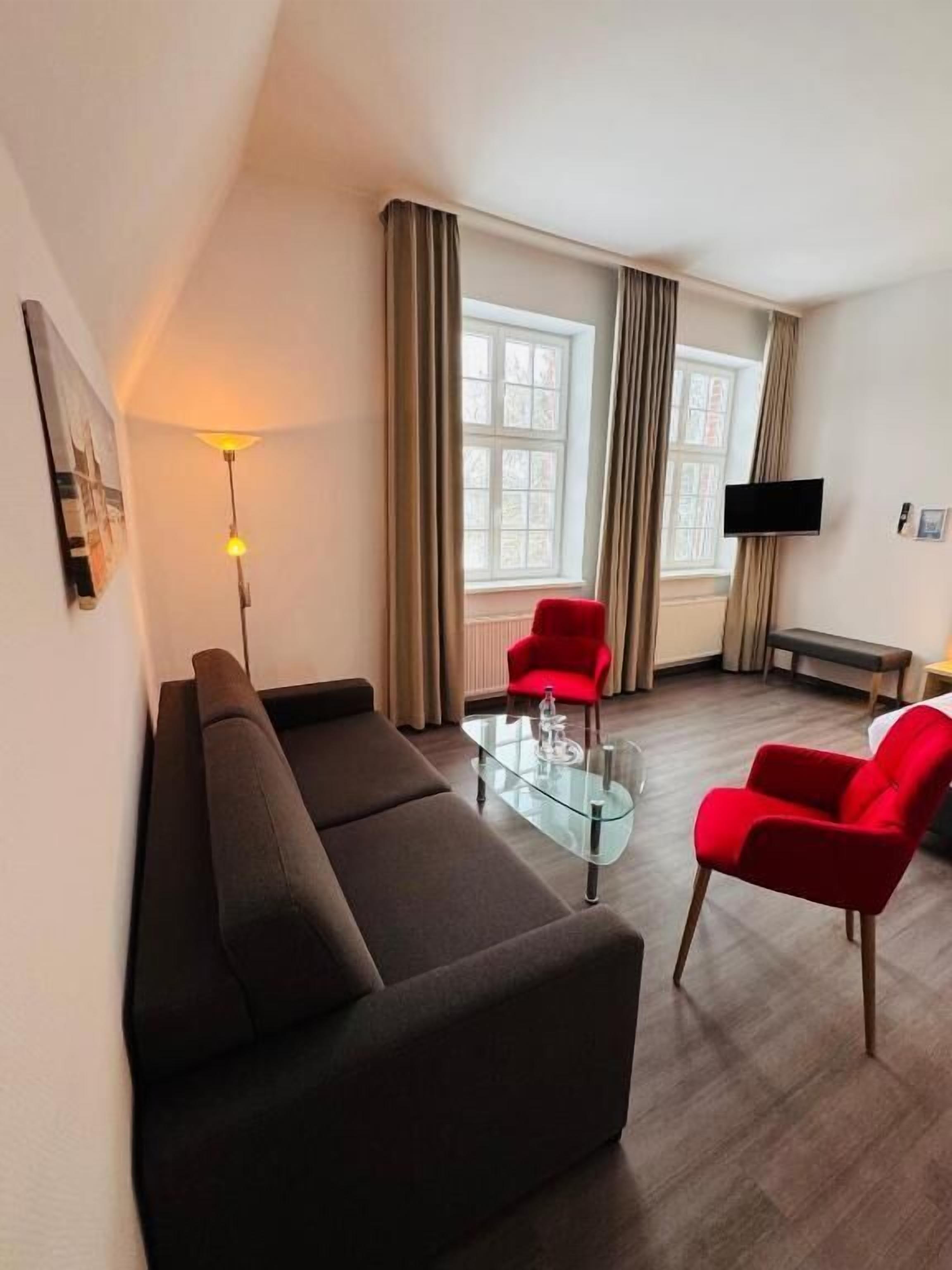 A living room with a sofa, two red chairs, a glass coffee table, a flat-screen TV, and a window with curtains.