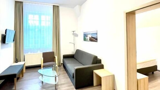 A modern hotel room with a sofa, a glass coffee table, a bench, a standing lamp, and a large window with curtains.