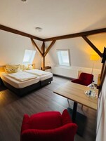 Una habitación con una cama, una silla roja, una mesa de madera y un ventanal.