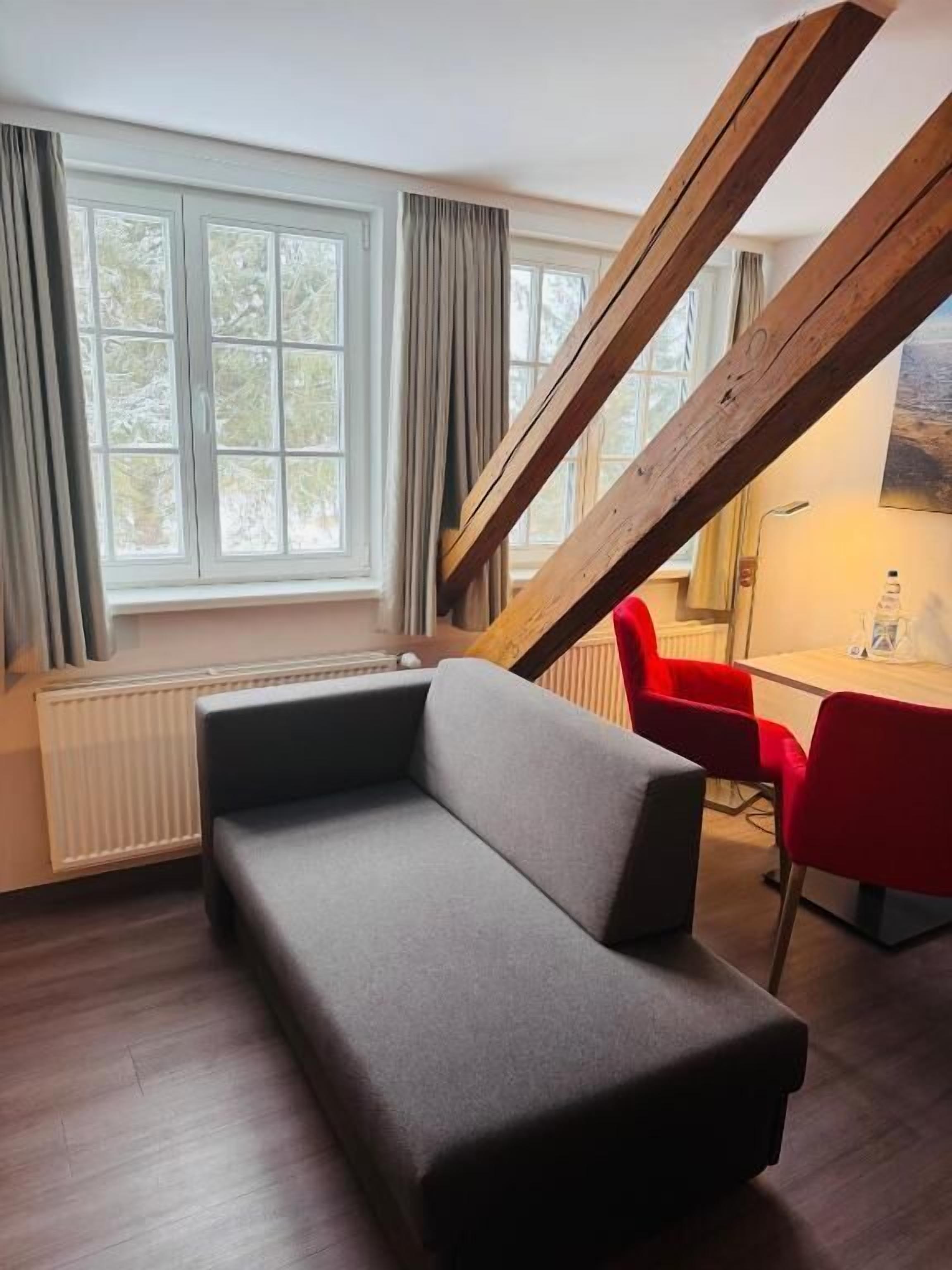 Una habitación con sofá gris, sillas rojas, una ventana con cortinas y estructura de vigas de madera.