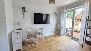 Una habitación con piso de madera, un escritorio blanco con cajones, una silla, un televisor montado en la pared y un ventanal corredero que da a un espacio exterior.