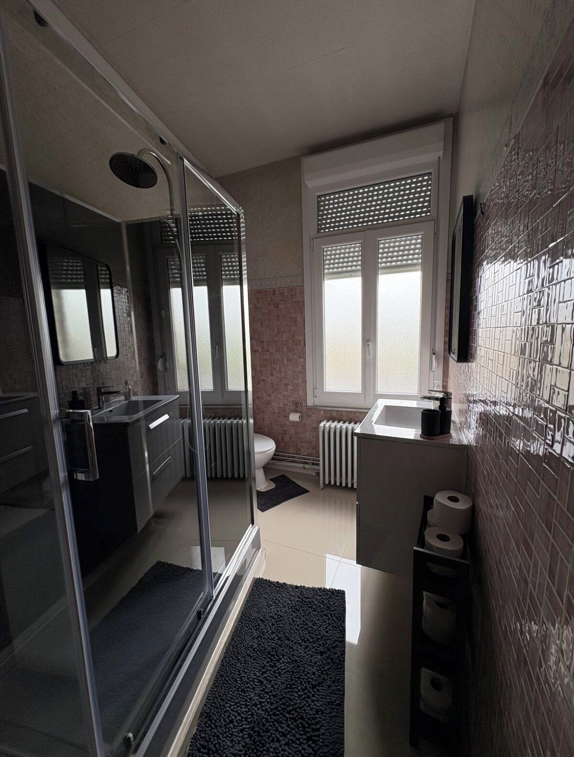 A modern bathroom with a glass shower enclosure, a toilet, a sink, and a window with blinds.