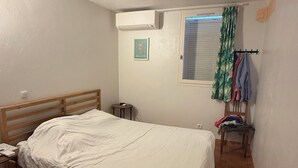 Une chambre avec un lit simple, une petite table de chevet, une chaise, une fenêtre avec des stores, un climatiseur fixé au mur et un rideau.