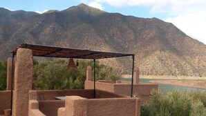 A rustic outdoor seating area with a metal roof, surrounded by mountains and a body of water.