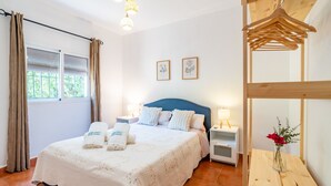 Un dormitorio con cama, cortinas, mesita de noche y un perchero, además de un jarrón con flores.