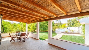 Un patio cubierto con suelo de baldosas, vigas de madera en el techo y vistas a un jardín y una piscina.