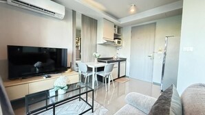 A modern living room with a flat-screen TV, a glass coffee table, a sofa, and a dining area with a white table and chairs. - 6th Avenue Surin by Win217 (Choeng Thale)