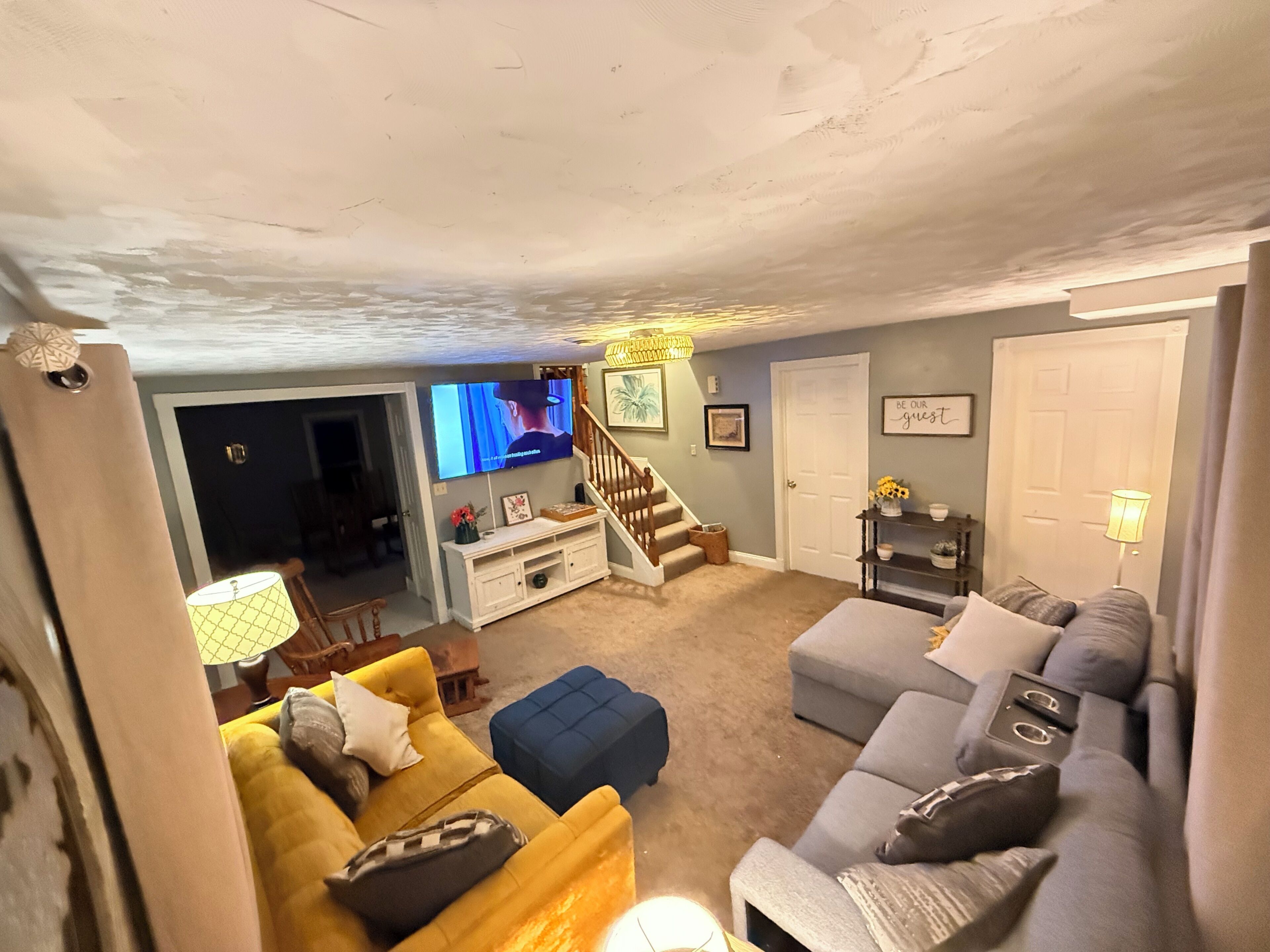 Ein Wohnzimmer mit einer Couch, Hocker, Fernseher, Treppe und einer Tür mit einem Schild.