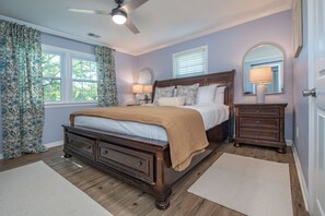 Une chambre à coucher comprenant un lit, des tables de chevet, une commode, un ventilateur de plafond et une fenêtre avec des rideaux.