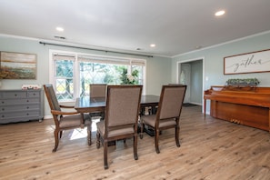 Un espace repas avec une table et des chaises en bois, un piano et une vue sur l’extérieur grâce à de grandes fenêtres.
