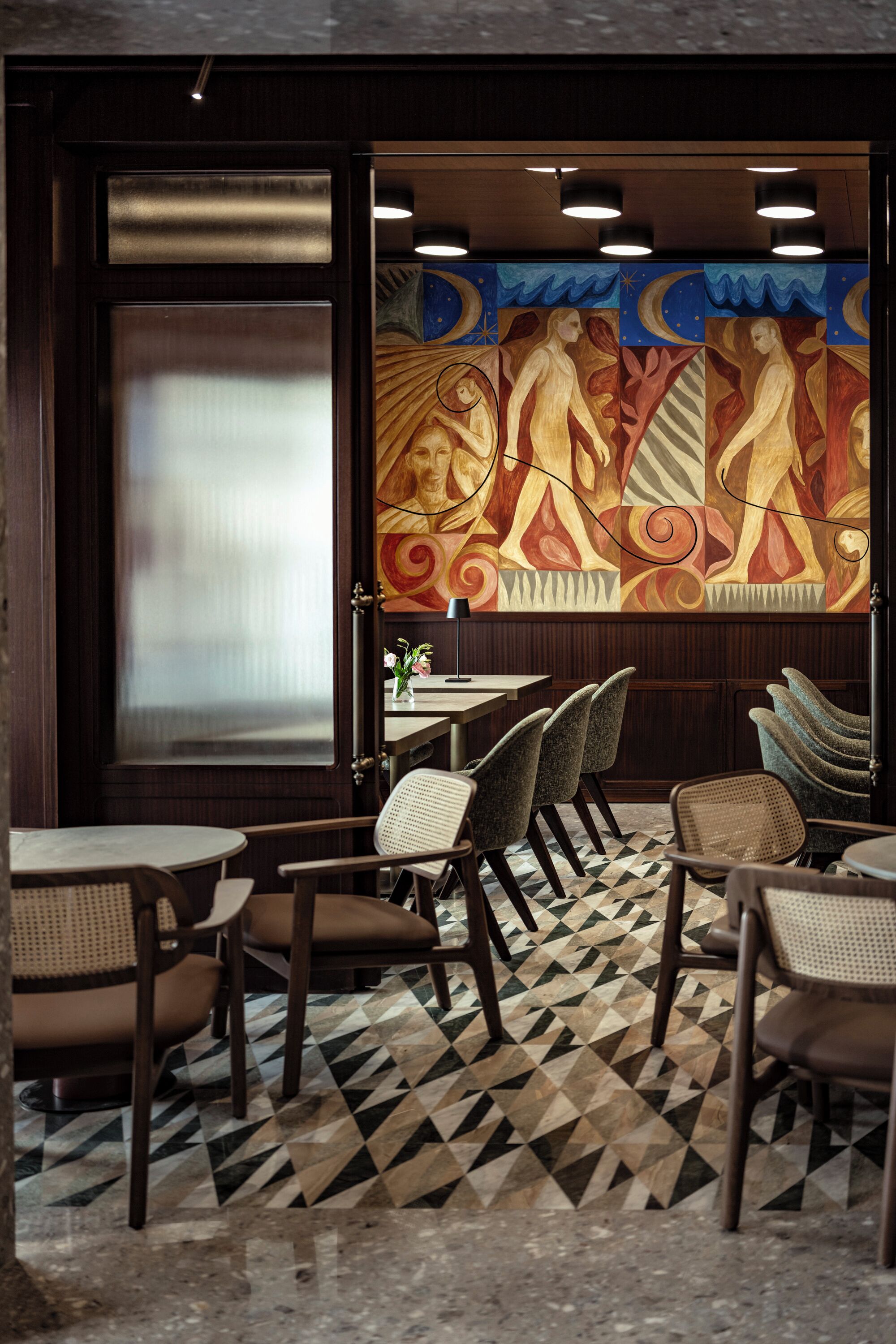 Interior de restaurante con suelo de patrón geométrico, sillas y mesas de madera, y un mural con figuras abstractas.