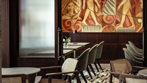 L’intérieur d’un restaurant présente un sol à motifs géométriques, des chaises et des tables en bois, ainsi qu’une fresque murale représentant des figures abstraites.