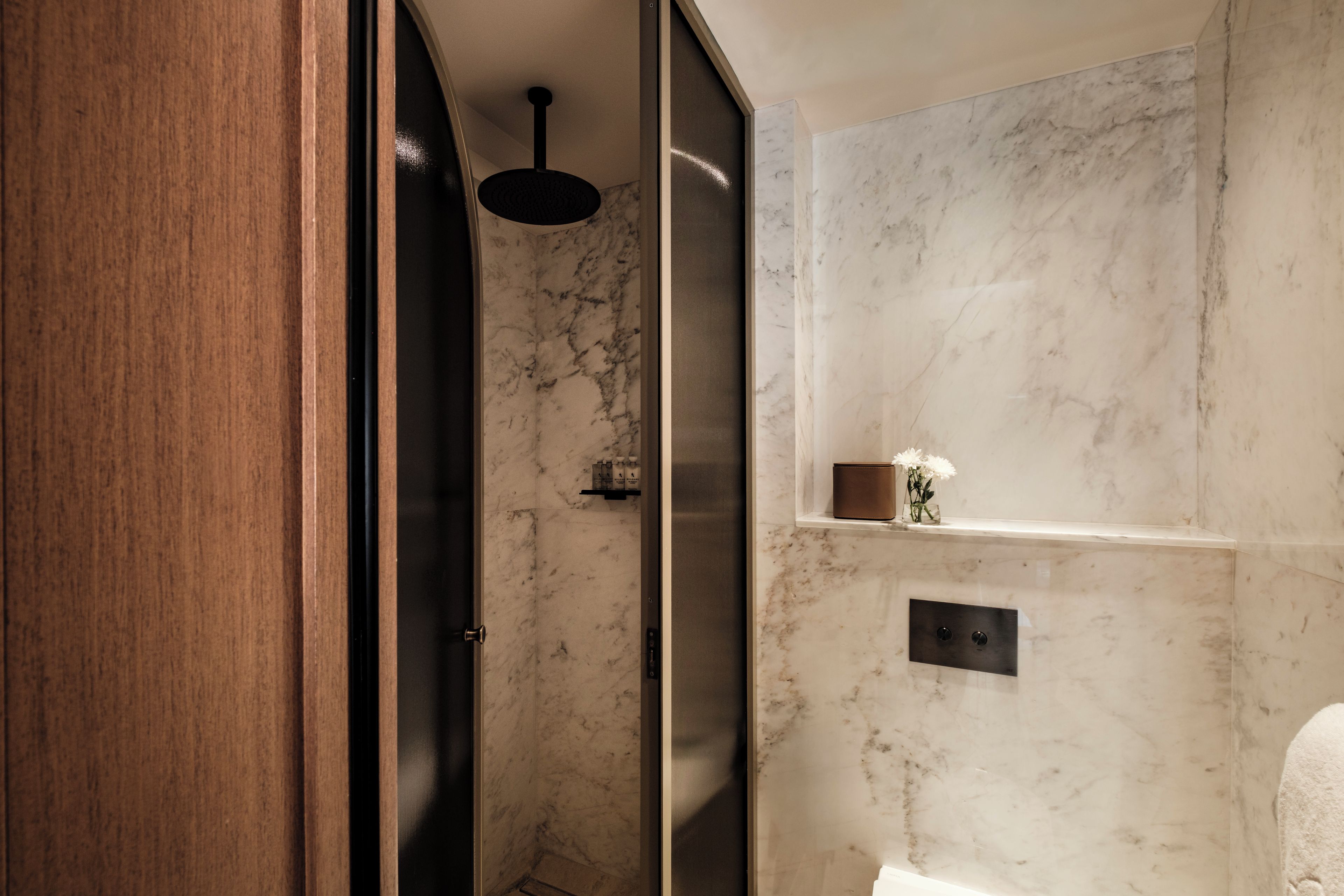 A modern bathroom with a wooden door, a shower with a black ceiling light, and a marble wall with a small shelf holding toiletries.