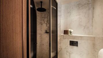 A modern bathroom with a wooden door, a shower with a black ceiling light, and a marble wall with a small shelf holding toiletries.