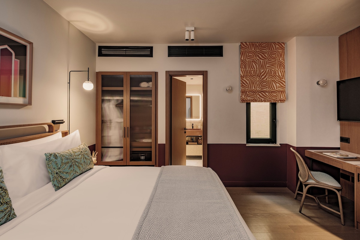 A hotel room with a bed, a desk, a chair, a wardrobe, and a bathroom visible through an open door.
