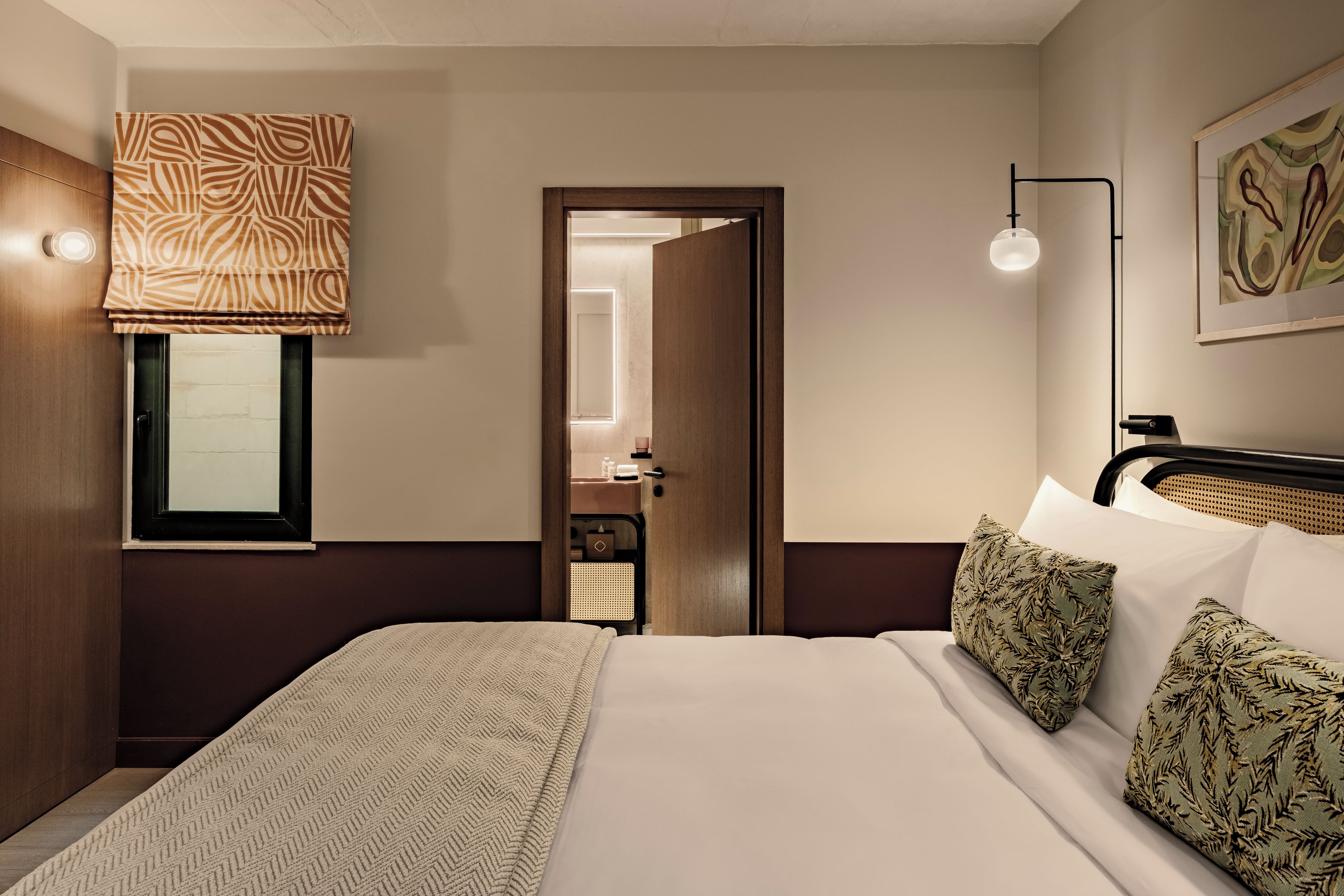 Una habitación de hotel con cama, ventana con persianas, mesita de noche, lámpara y un cuadro en la pared.