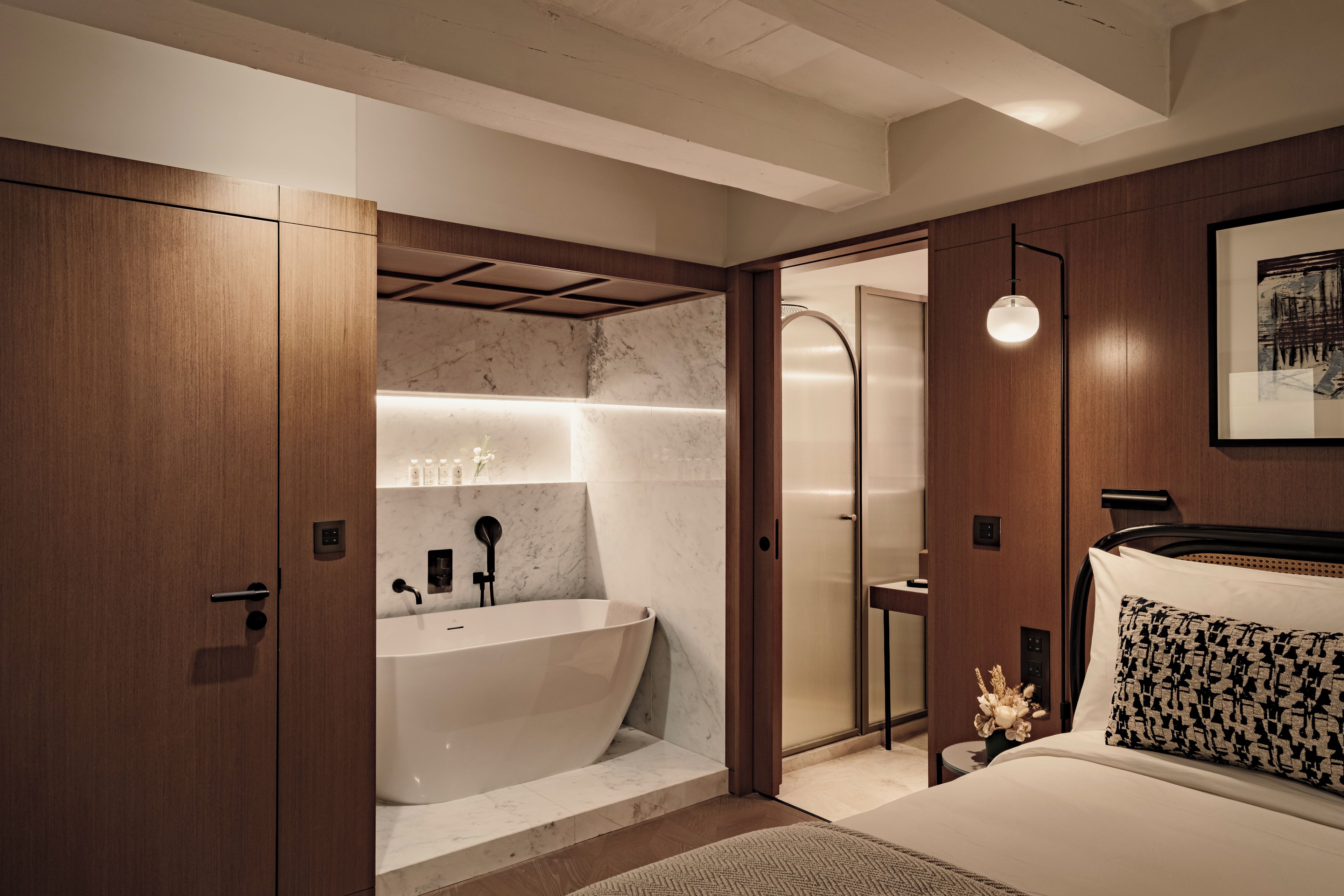 A modern bathroom with a freestanding bathtub, a shower area, and a bed with a patterned pillow.