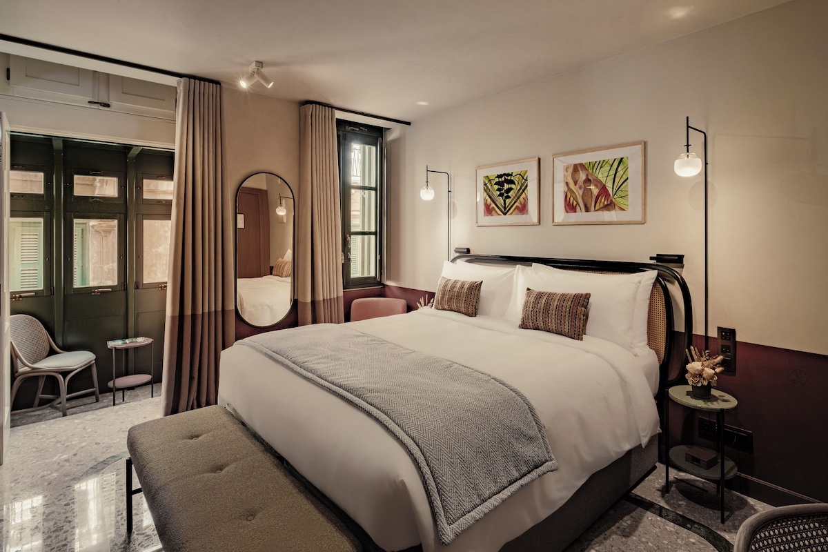 A modern hotel room with a large bed, a sitting area with a chair, a small table, and a mirror. There are two framed artworks on the wall.