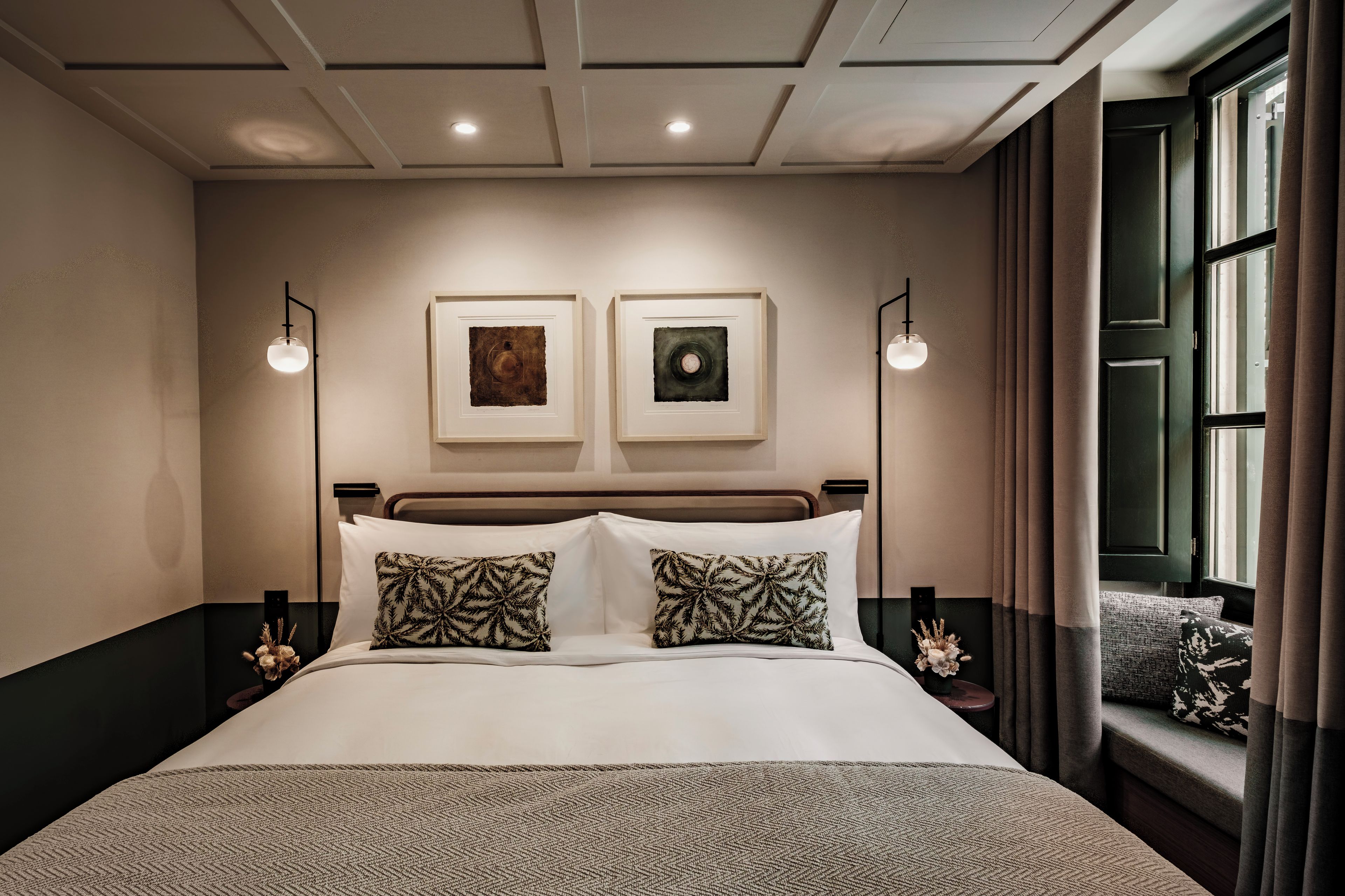Une chambre d’hôtel avec un grand lit, deux œuvres d’art encadrées au mur, une fenêtre avec des rideaux et une table de chevet avec une lampe.