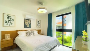 Hotellihuone, jossa on sänky, yöpöytä, ikkuna verhoilla ja seinätaidetta.