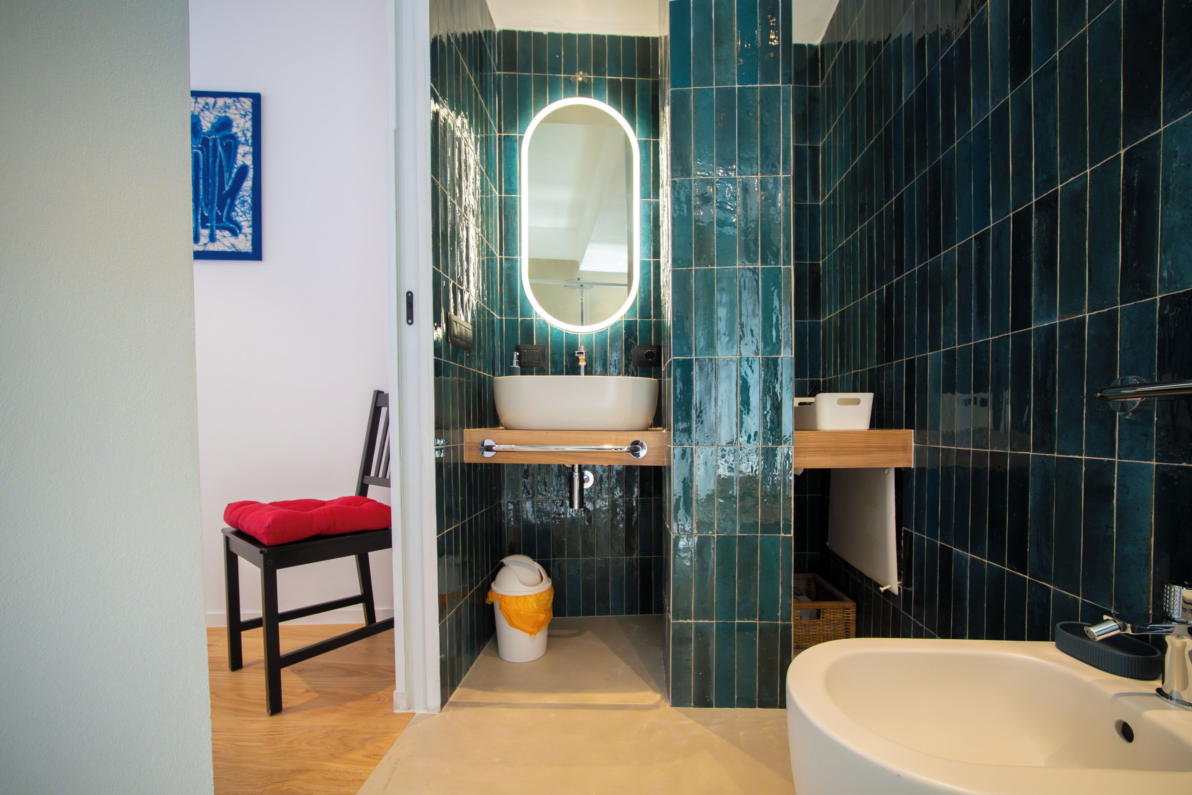A bathroom with a white sink, a mirror with a light, a red chair, and a trash bin.