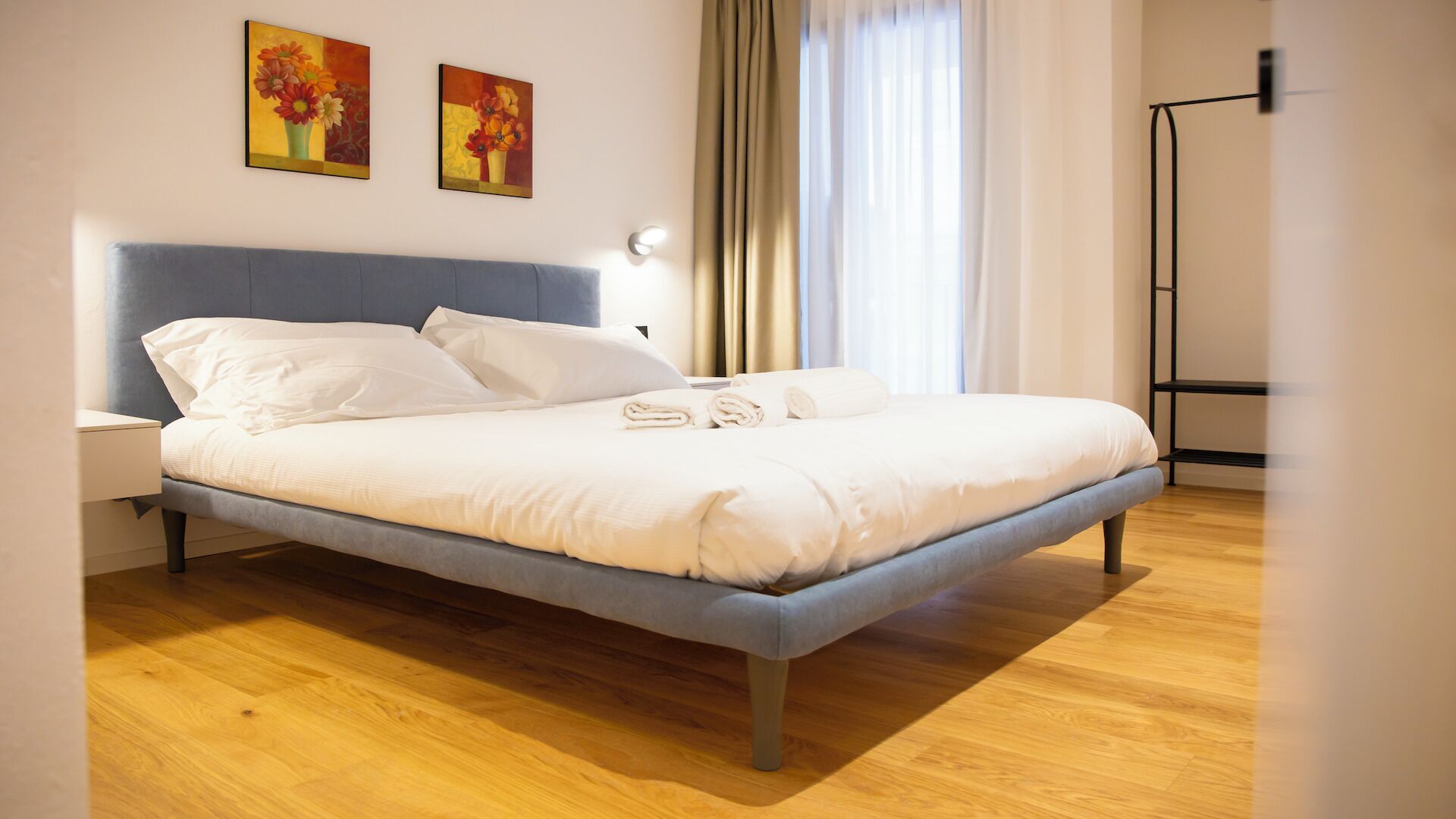 Un lit bien fait, avec du linge de lit blanc, une tête de lit grise et un plancher en bois. Deux tableaux sont accrochés au mur au-dessus du lit, et une lampe de sol est visible sur la droite.