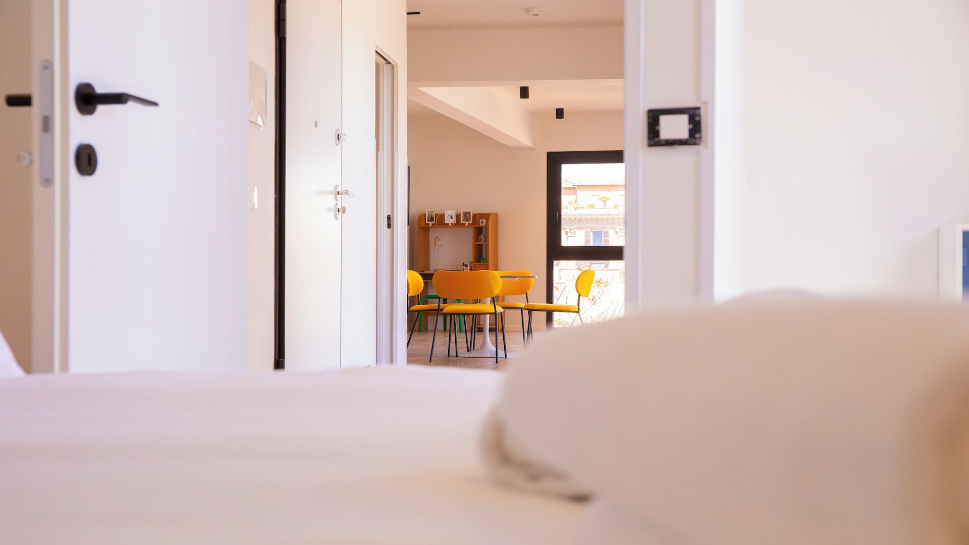 A hotel room with a bed, a door, and a view of a dining area with yellow chairs.