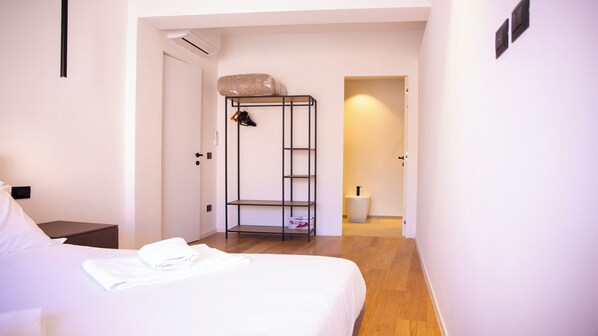 A hotel room with a bed, a small bedside table, a wall-mounted shelf, and a door leading to a bathroom.