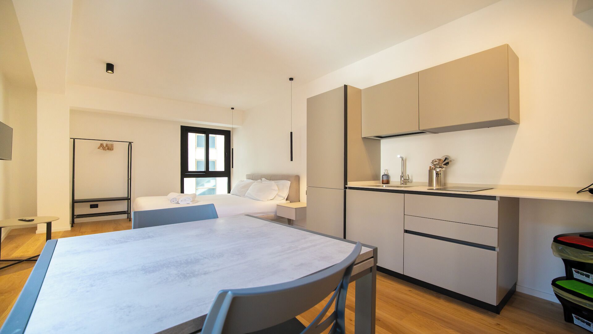 Ein modernes Hotelzimmer mit Esstisch, Küchenzeile, einem Bett und einem großen Fenster.