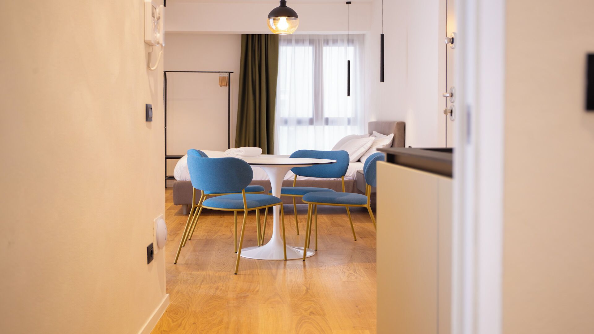 A modern hotel room with a dining area featuring a round table and blue chairs, a bed with white linens, and a large window with curtains.