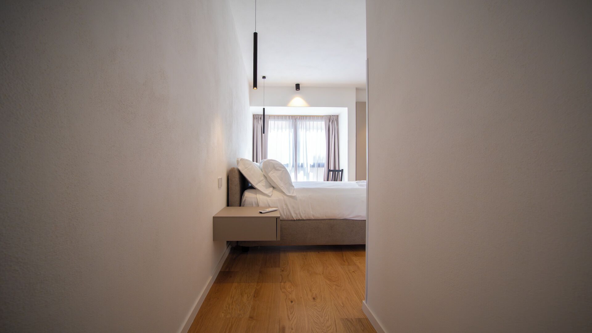 Ein schmaler Gang führt zu einem Schlafzimmer mit Bett, Nachttisch und einem Fenster mit Vorhängen.