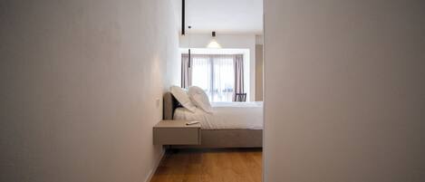Ein schmaler Gang führt zu einem Schlafzimmer mit Bett, Nachttisch und einem Fenster mit Vorhängen.