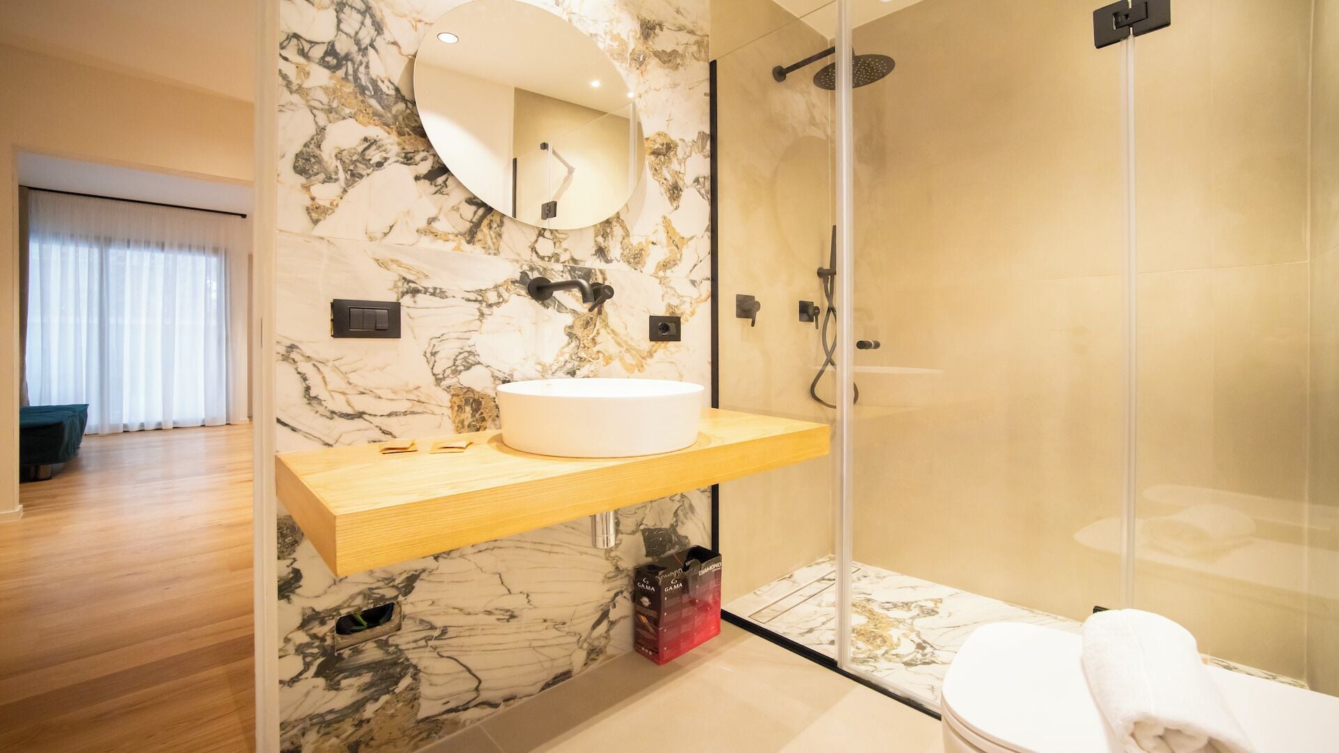 Un baño moderno con pared de mármol, espejo redondo, tocador de madera, cabina de ducha de vidrio y un inodoro.
