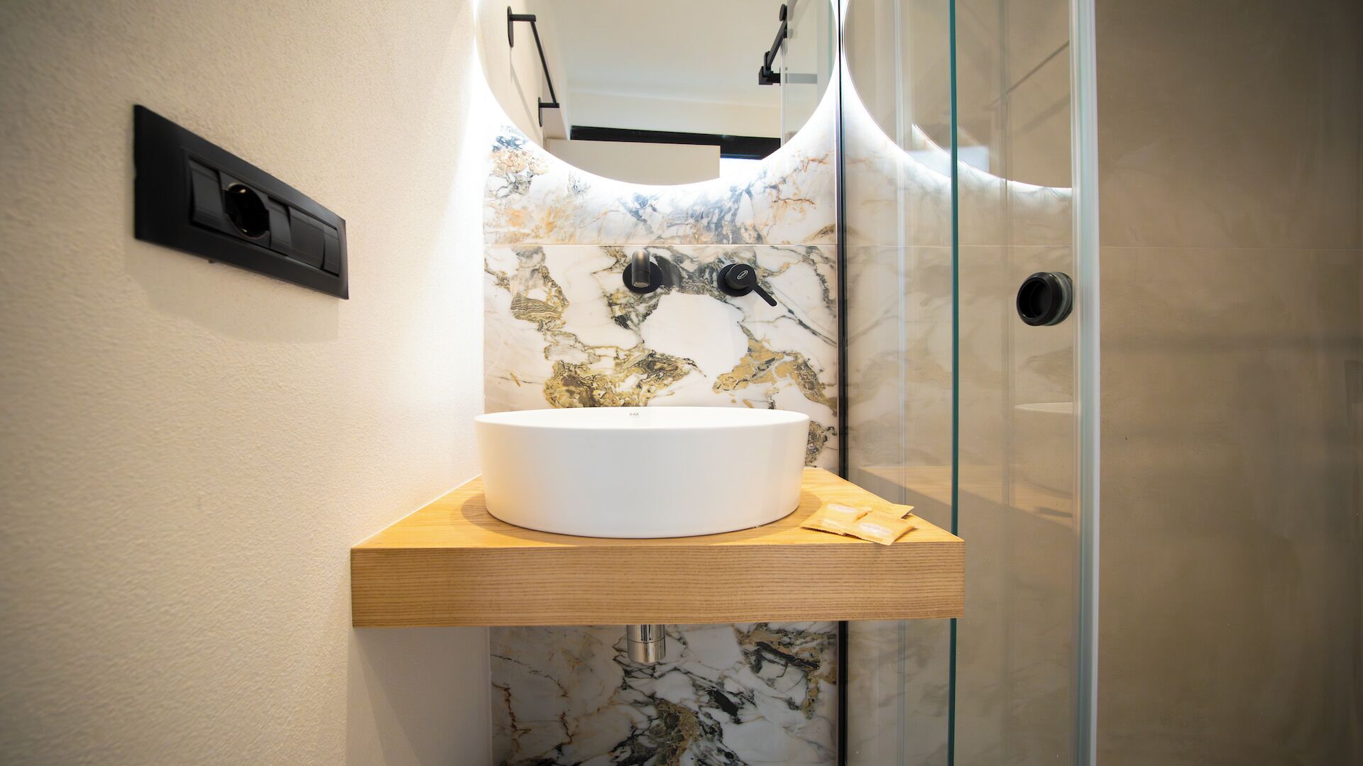 Une salle de bain avec un lavabo blanc posé sur une étagère en bois, un pare-baignoire en marbre et une douche à parois de verre.