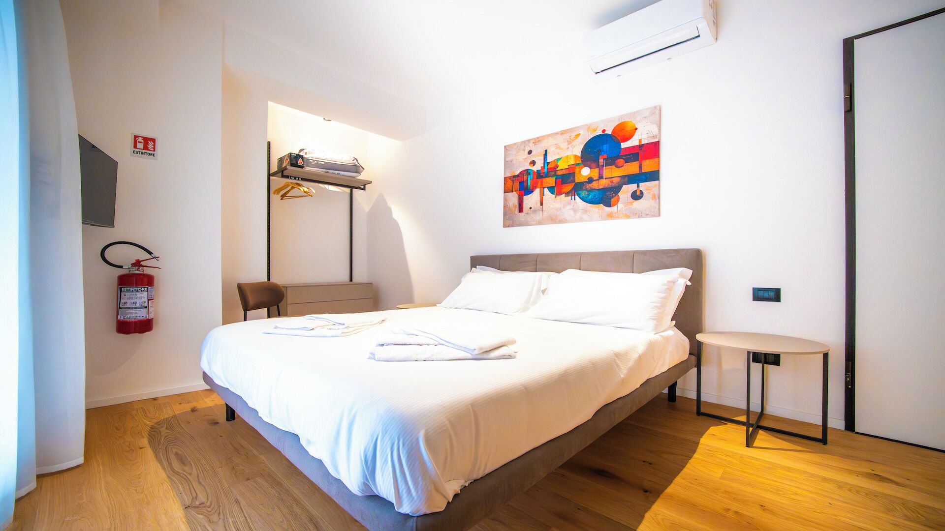 Quarto de hotel moderno com uma cama, uma pequena mesa redonda, um quadro na parede e um extintor de incêndio.