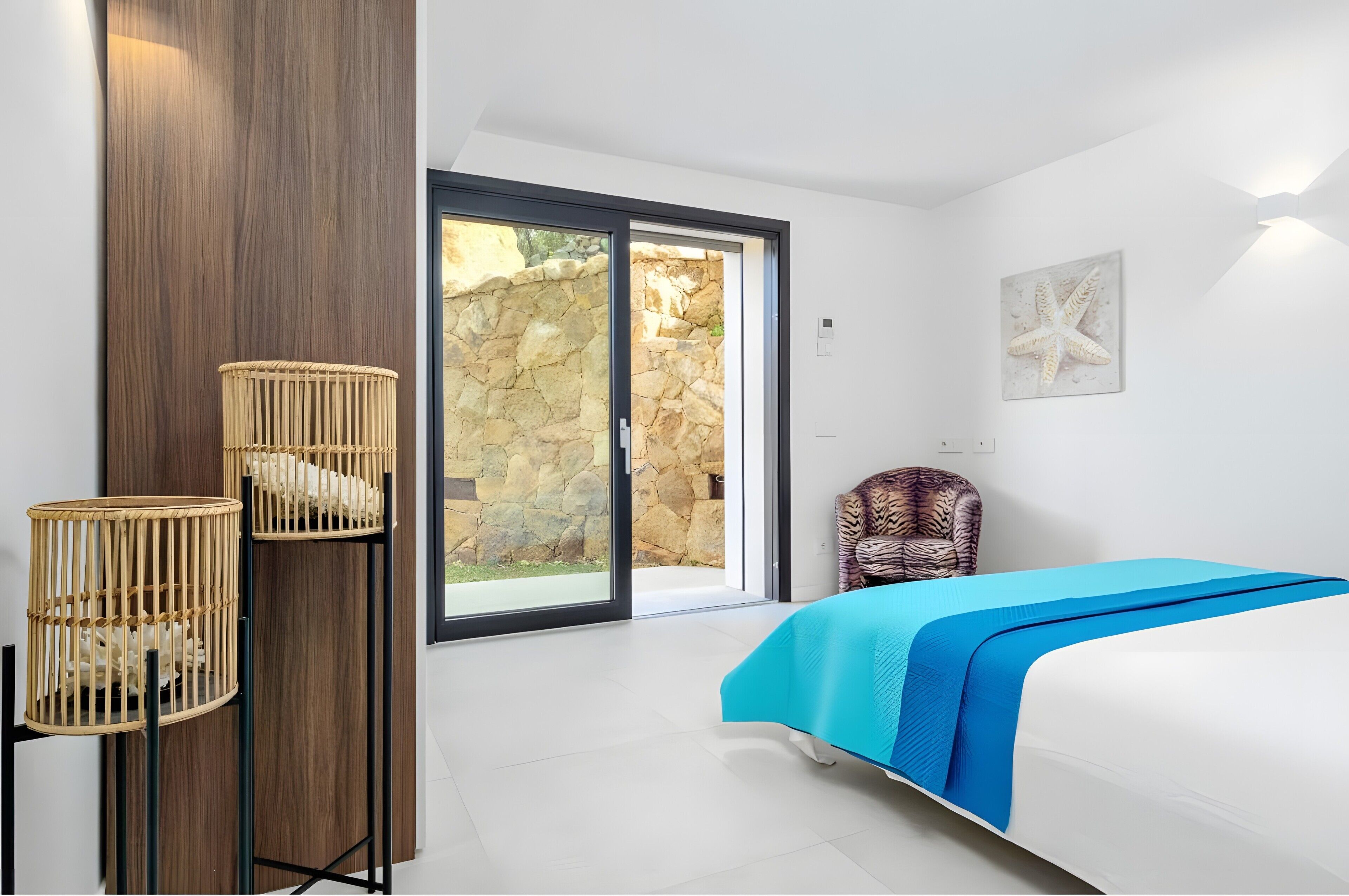 Ein Schlafzimmer mit einem Bett, einem Sessel, einer Wandlampe und Blick durch eine Glastür.