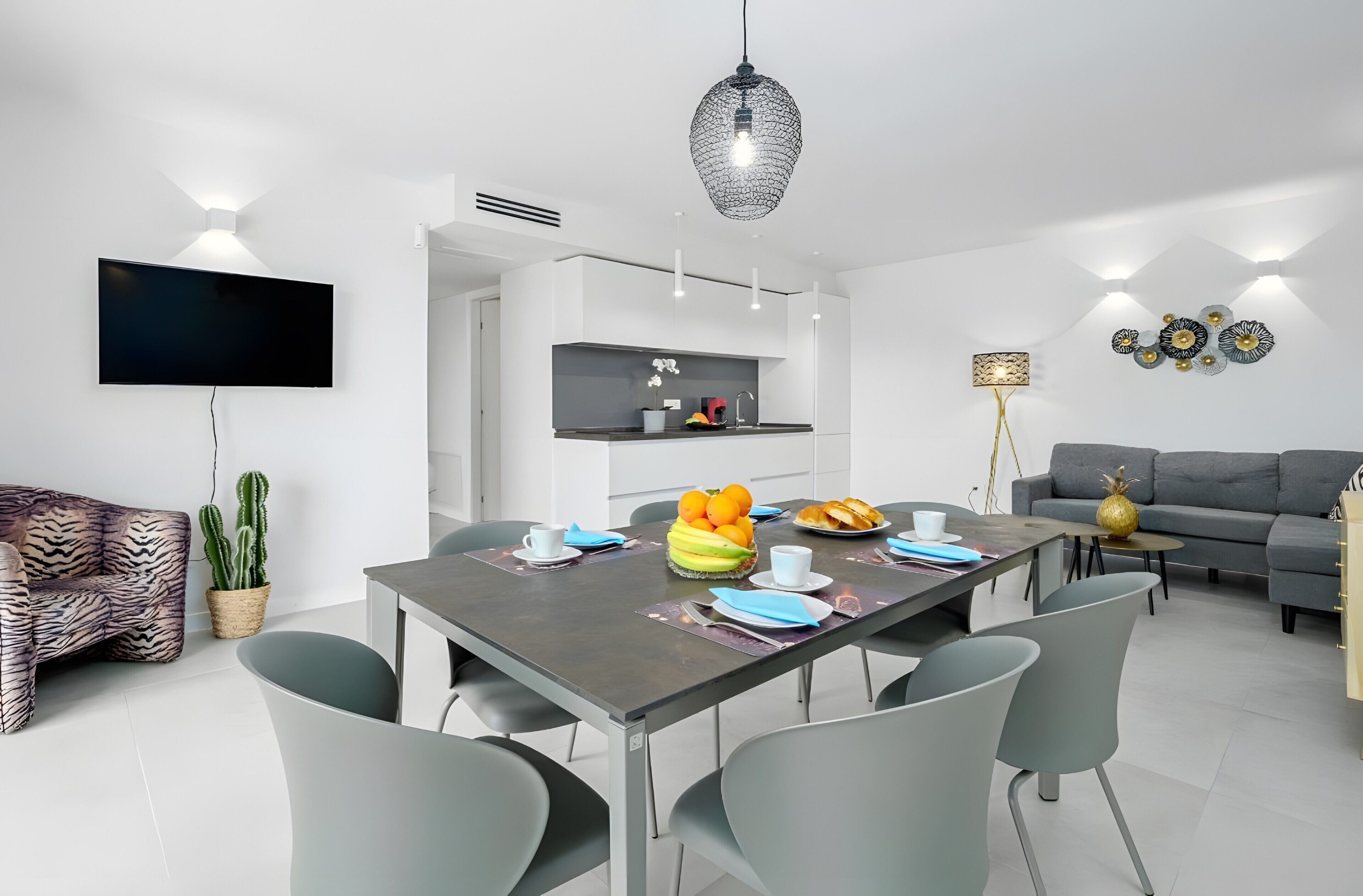 Un salon moderne doté d’un téléviseur à écran plat, d’un coin repas avec une table dressée pour deux personnes et d’un canapé gris.