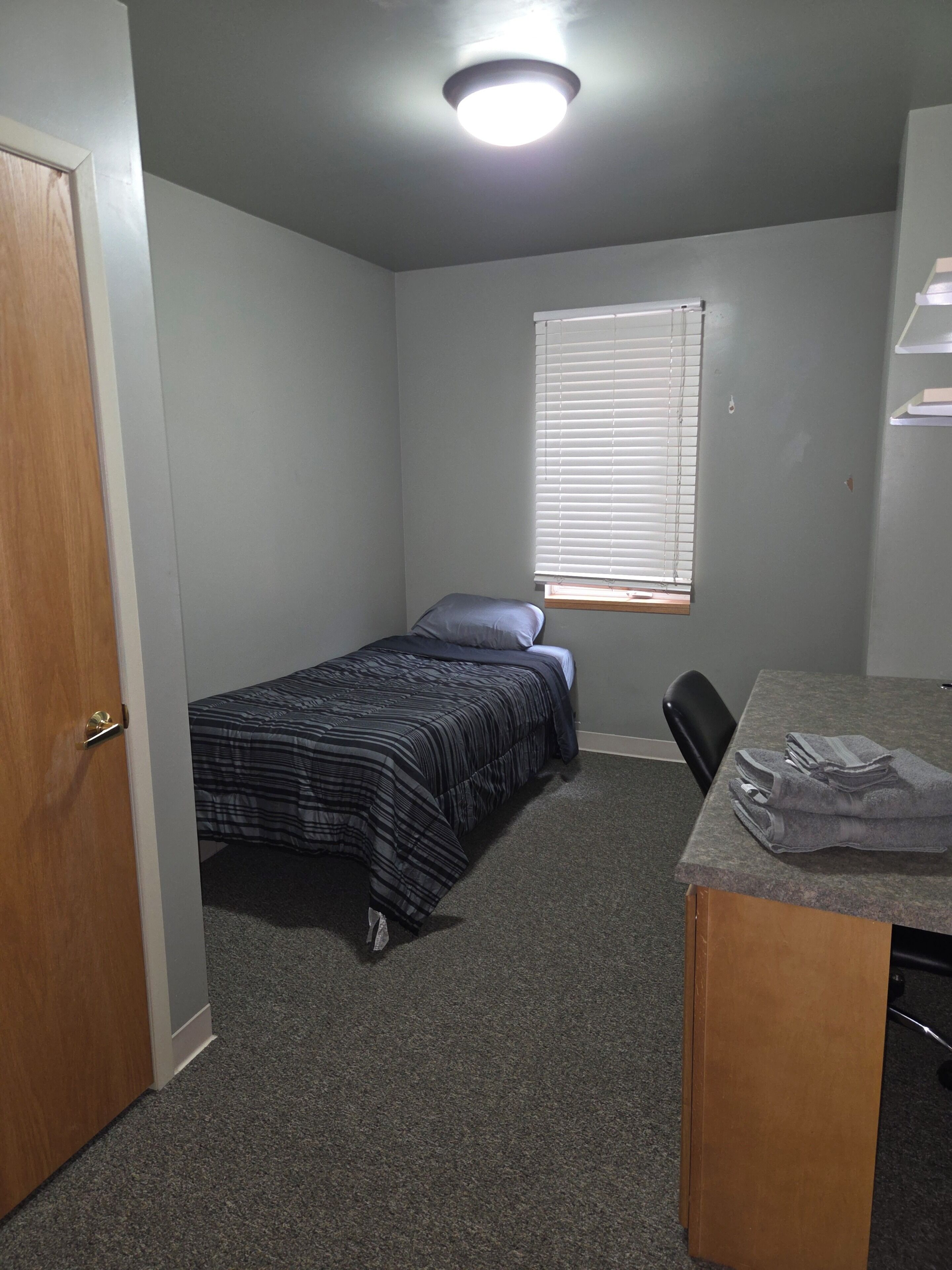 A small, single-bed room with a desk and chair, a window with blinds, and a door.