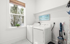 Une buanderie avec deux machines à laver blanches, une fenêtre donnant sur des arbres, une carte accrochée au mur et un aspirateur.