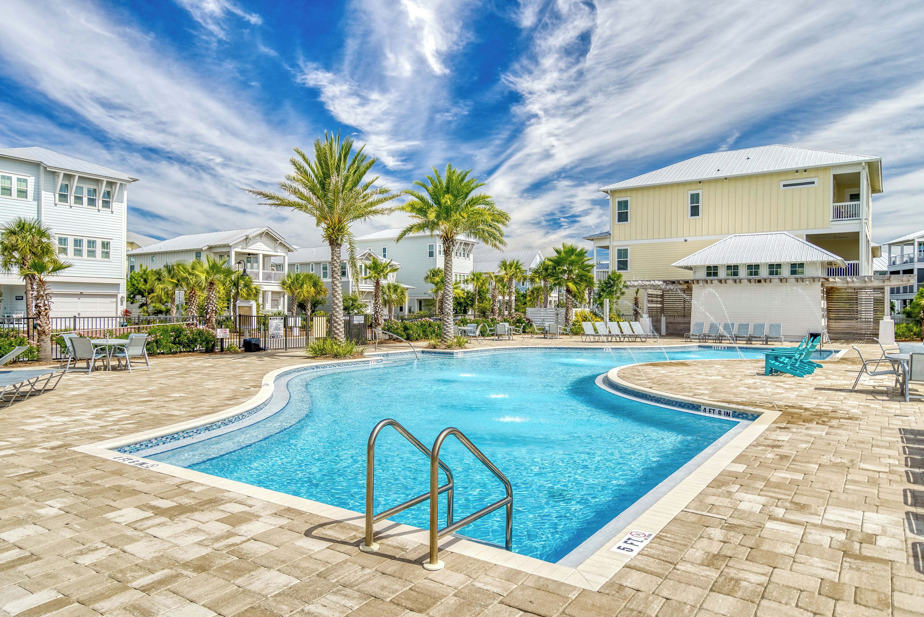 A swimming pool with a tiled deck, palm trees, and houses in the background.