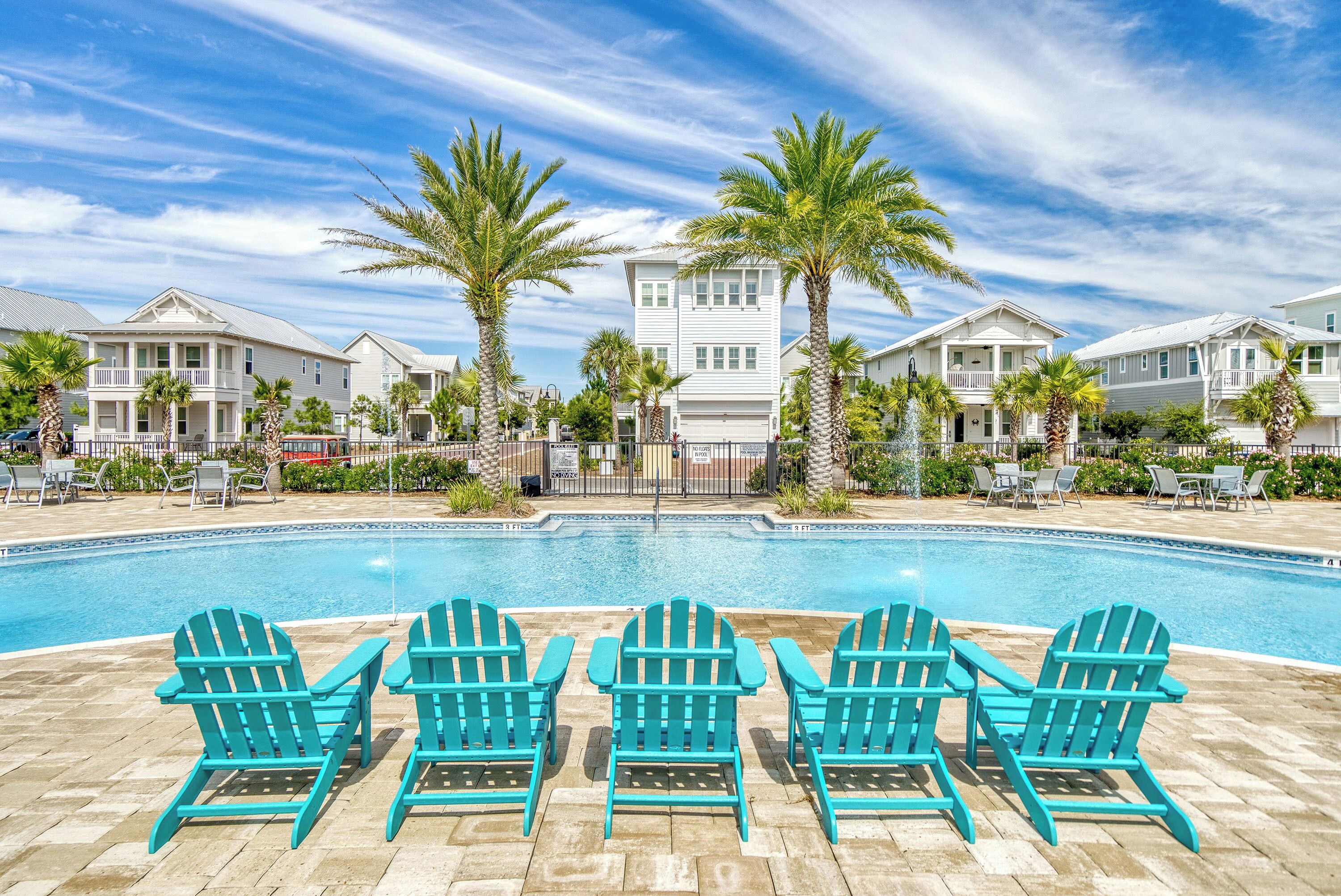 Une piscine bordée de chaises Adirondack turquoise, de palmiers et d’une rangée de maisons blanches.