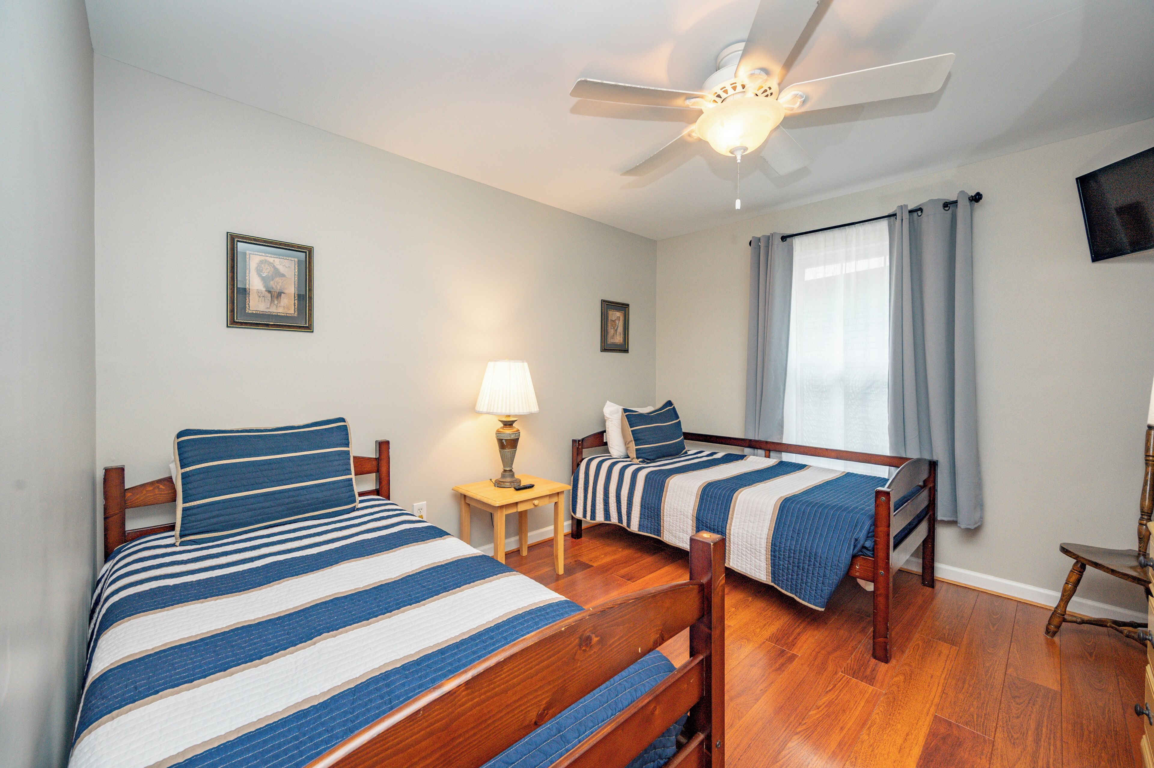 A bedroom with two beds, a ceiling fan, a small table with a lamp, a chair, and a television on the wall.