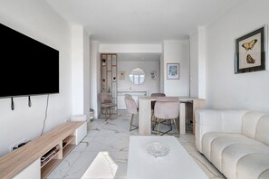 Ein modernes Wohnzimmer mit Flachbildfernseher, einem weißen Sofa, einer Essecke mit Tisch und Stühlen sowie einer Küche im Hintergrund.
