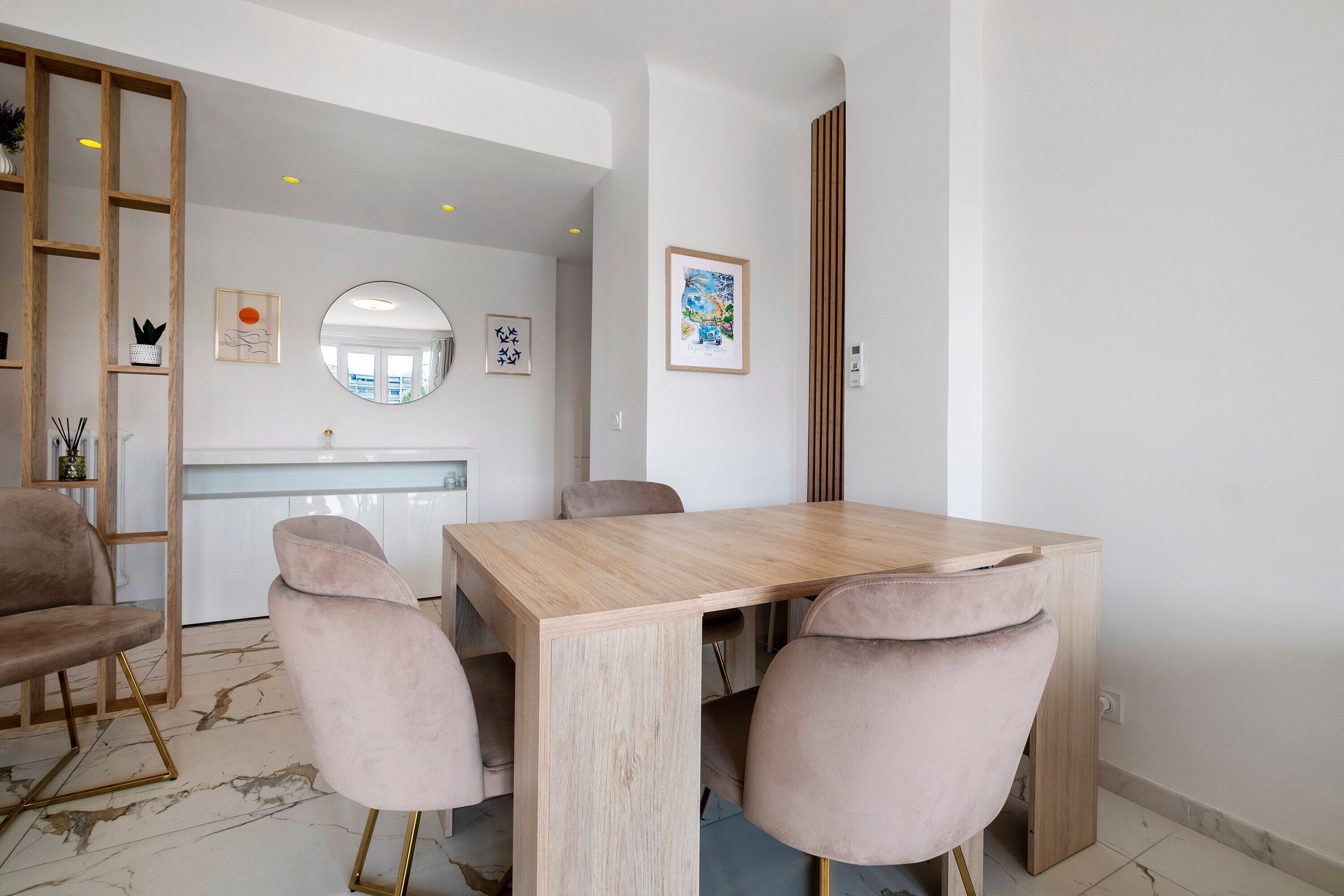 A modern dining area with a wooden table and four chairs, a kitchen in the background, and a mirror on the wall.