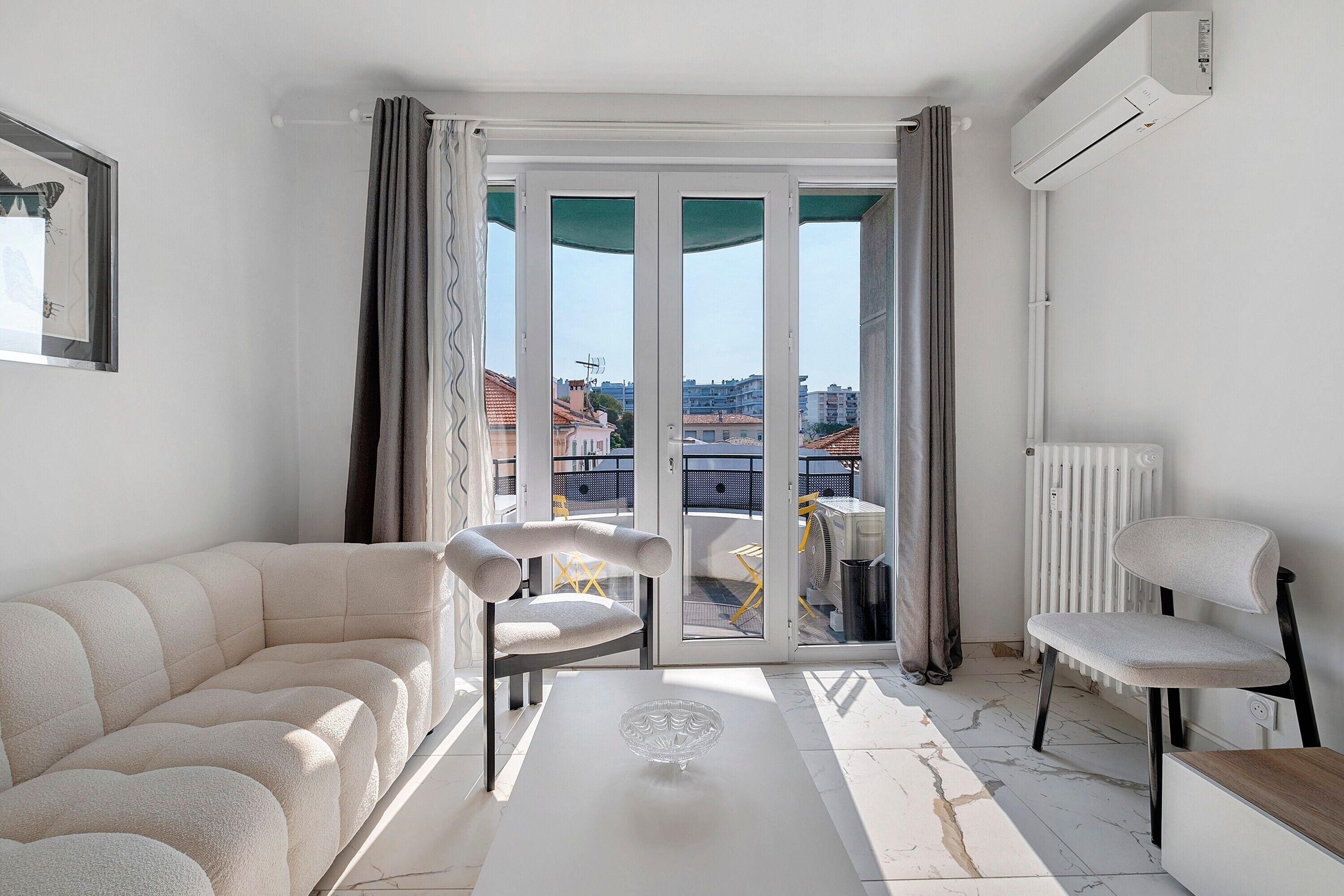 Un salon moderne avec un canapé, un fauteuil, une table basse et une vue sur l’extérieur, visible à travers une porte vitrée.