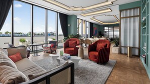 Habitación de hotel moderna con ventanas amplias, un sofá, dos sillones rojos, una mesita de centro y un comedor.