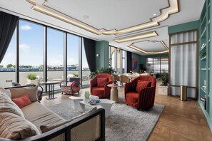 Ein modernes Hotelzimmer mit großen Fenstern, einem Sofa, zwei roten Sessel, einem Couchtisch und einem Essbereich.