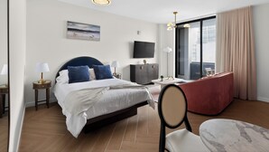 Habitación de hotel con cama, mesitas de noche, televisor, sofá rojo, una silla y un ventanal con cortinas.